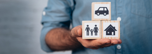 panoramic crop of man holding wooden cubes with car, family and house  - Photo, Image