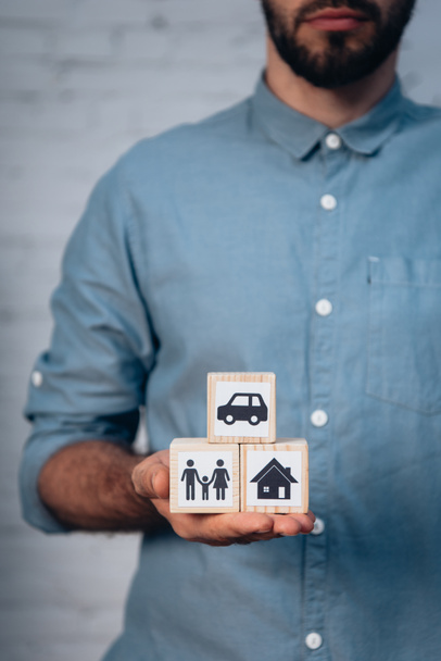 vista recortada del hombre barbudo sosteniendo cubos de madera con coche, familia y casa en la mano - Foto, Imagen