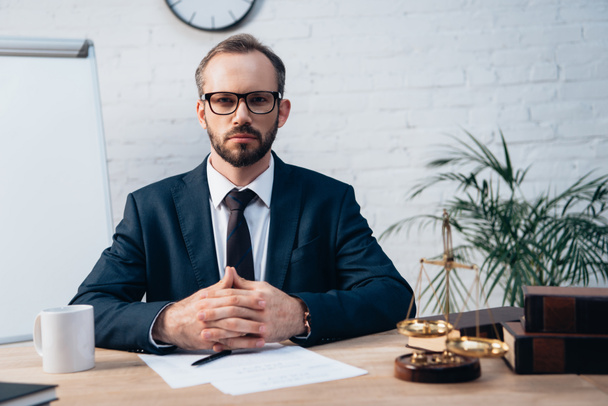 bearded lawyer in glasses looking at camera and sitting with clenched hands at desk - Foto, imagen