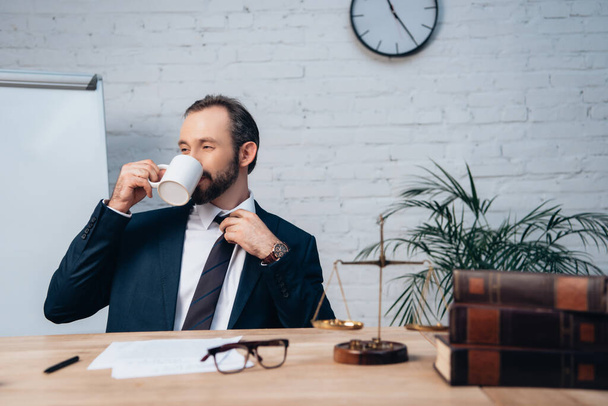 barbudo abogado en traje tocando corbata mientras bebiendo café en oficina - Foto, imagen