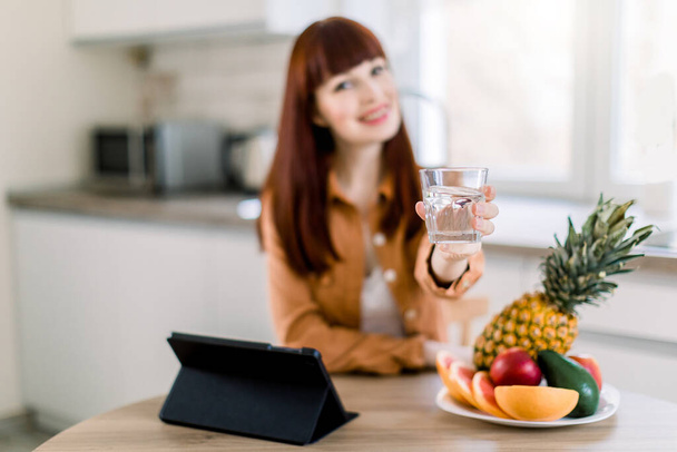 Beautiful young woman with glass of water in kitchen, sitting at the table and showing it to camera with nice smile, while having healthy fruit breakfast and working on tablet. Focus on glass - Photo, image