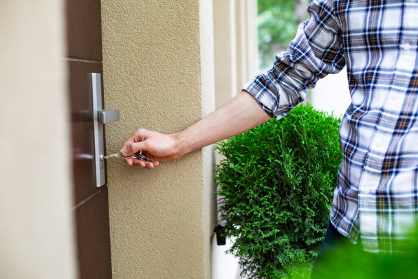 A man in casual clothes, opening, closing the front door of the house on a sunny day. Male hand holding a bunch of keys at an elegant house entrance. Focus on the hand inserting the key into the lock. - Foto, Imagen