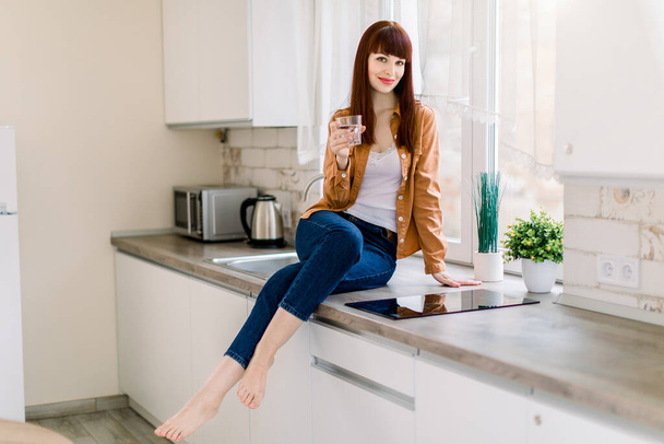 Attractive young woman in denim jeans and mustard casual shirt, sitting with glass of drinking water on the kitchen countertop in modern light home kitchen. Healthy lifestyle concept - Foto, Bild