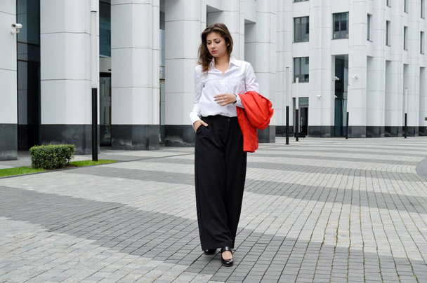 近代的なオフィスの背景に美しいビジネス女性。ビジネスセンターの近くで彼の手の中に赤いジャケットと白いシャツと黒いズボンの暗い髪の女の子 - 写真・画像