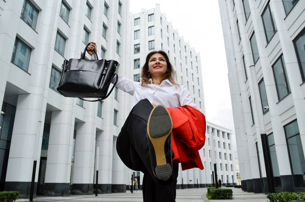 Beautiful business woman on the background of the modern office. A girl with dark hair in a white shirt and black trousers with a red jacket and a black bag in her hands. Photo bottom up - Fotó, kép