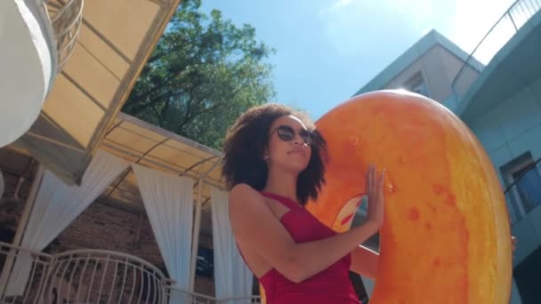 Mujer americana de pelo rizado con estilo enfocado en gafas y paseos de traje de baño rojo desde la habitación del hotel hasta el fondo de la piscina de la playa de rayos de sol, lleva la celebración de anillo inflable boya de vida naranja, vista inferior - Imágenes, Vídeo