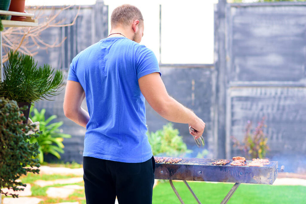 Knappe atleet man koken barbecue buiten. Brutale man met een baard, staande en gekookt vlees op de binnenplaats. Achteraanzicht. Kopieerruimte. - Foto, afbeelding