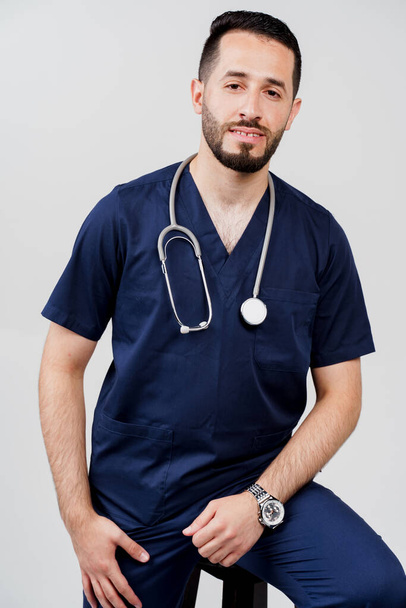 Arab orvostanhallgató sztetoszkóppal, sebészeti egyenruhában. Jóképű szakállas férfi a stúdióban. Magabiztos orvos konzultáción - Fotó, kép