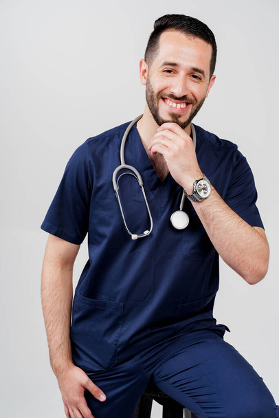 Estudiante árabe de medicina con estetoscopio en uniforme quirúrgico sonríe con fondo en blanco. Hombre barbudo guapo en el estudio. Médico de confianza en consulta - Foto, imagen