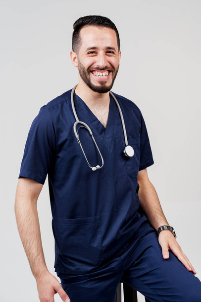 Arabian medical student with stethoscope in surgical uniform smiles on blanked background. Handsome bearded arab man in studio. Confident doctor at consultation - Photo, Image