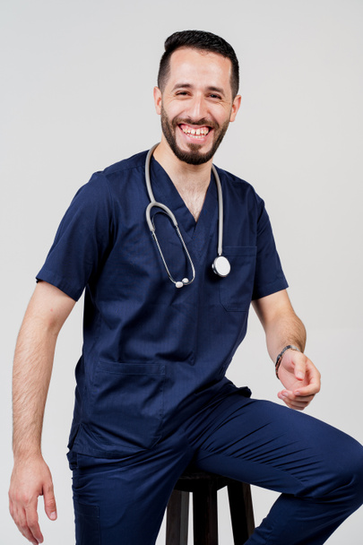 Arab orvostanhallgató sztetoszkóppal, sebészeti egyenruhában, mosollyal, üres háttérrel. Jóképű szakállas férfi a stúdióban. Magabiztos orvos konzultáción - Fotó, kép