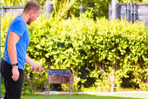 Красивый спортсмен готовит барбекю на открытом воздухе. Жестокий бородатый мужчина стоял и готовил мясо во дворе. Вид сзади. Копирование пространства. - Фото, изображение