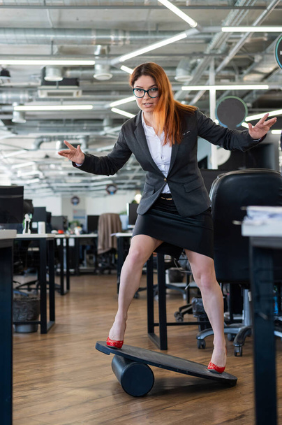 A woman in a suit is engaged on a rocker-skateboard during a break at work. A female office employee in high heels stands on a balance board. - 写真・画像