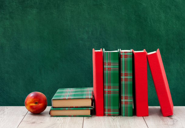 Zpátky do školy, hromada knih v barevných obalech a červená broskev na dřevěném stole s prázdným zeleným školním pozadím. Dálkové domácí vzdělání. Karanténní koncept pobytu doma - Fotografie, Obrázek