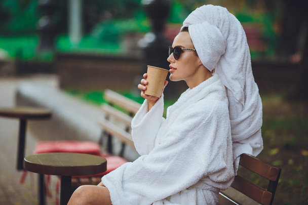 Une fille en peignoir et une serviette sur sa tête est assise en train de boire du café - Photo, image
