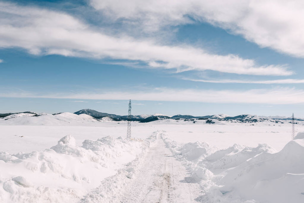 Υψηλή πόλους γραμμή ισχύος σε μια χιονισμένη πεδιάδα κοντά σε ένα δρόμο με βουνά στον ορίζοντα. - Φωτογραφία, εικόνα