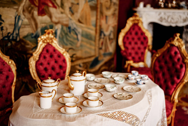 Um conjunto de chá para quatro em uma mesa com toalha de mesa de renda e cadeiras com aro de ouro e guarnição vermelha. - Foto, Imagem
