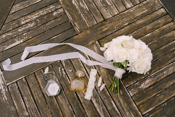 Bouquet di rose bianche con un lungo nastro in sviluppo per la sposa con giarrettiera, bottiglia di profumo, anello nuziale, orecchini e spighette su vecchie tavole di legno. - Foto, immagini