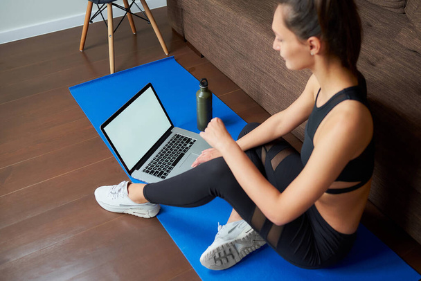 A sideways photo of a sporty girl in black tight pants and top is sitting on the blue yoga mat with a computer and a bottle at home. A mockup of a laptop and fitness woman. - Photo, Image