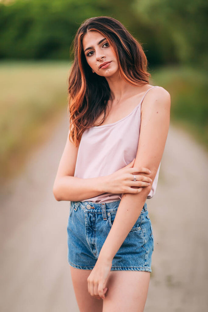 Portrait of beautiful sexy young woman with long brown hair in pink tank top and denim shorts posing outdoors at summer sunset, sensual, serious, blurry nature background - Foto, Imagen