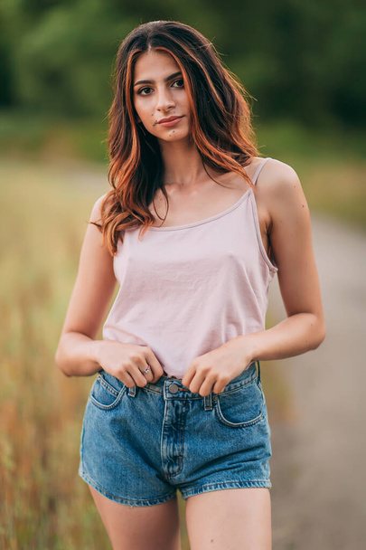 Portrait of beautiful sexy young woman with long brown hair in pink tank top and denim shorts posing outdoors at summer sunset, sensual, serious, blurry nature background - Foto, imagen