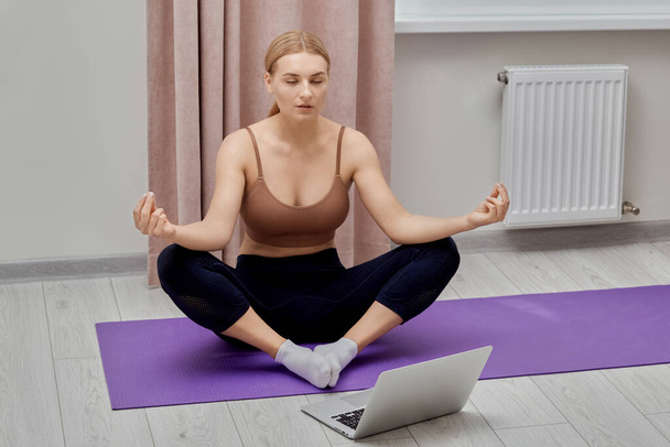 A beautiful young girl is engaged in yoga and looks into the laptop screen. - Foto, Imagen