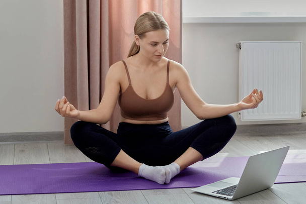 A beautiful young girl is engaged in yoga and looks into the laptop screen. - Photo, Image