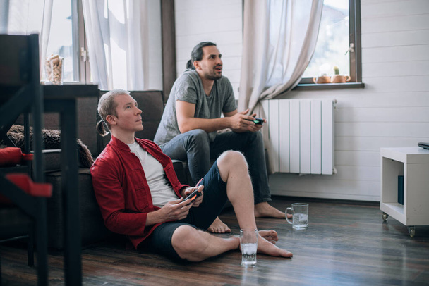 Ребята играют в видеоигры на диване. Два молодых человека с джойстиками в руках с энтузиазмом играют в игру на консоли - Фото, изображение