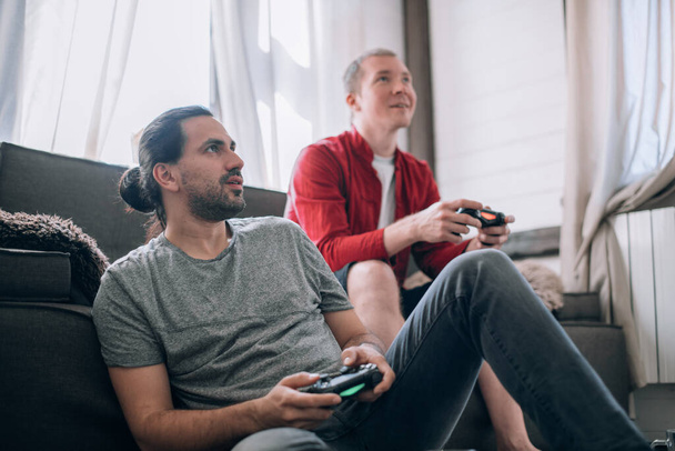 男たちはソファでビデオゲームをしている。両手にジョイスティックを持つ2人の若い男が熱狂的にコンソール上でゲームをプレイしています - 写真・画像