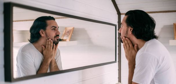 Een knappe man kijkt in de spiegel. Persoonlijke verzorging van mannen. Een jongeman onderzoekt zichzelf in de spiegel in de slaapkamer. - Foto, afbeelding