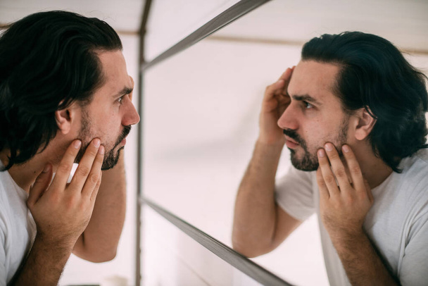 Un hombre guapo se mira en el espejo. El cuidado personal de los hombres. Un joven se examina a sí mismo en el espejo del dormitorio. - Foto, Imagen