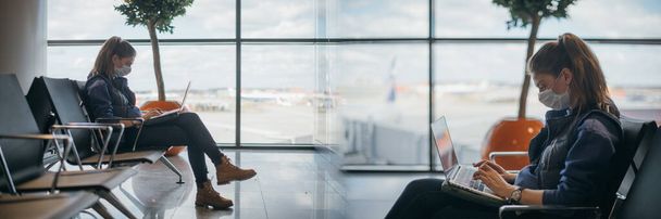 Una mujer está sentada en el aeropuerto con un portátil en una máscara médica. Una joven está esperando la salida en la puerta, trabajando en línea. Responsabilidad social, protección contra el virus. - Foto, imagen