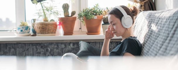 Egy lány ül a kanapén egy telefonnal a kezében. Egy fiatal nő otthon pihen, zenét hallgat fejhallgatón, videókat néz okostelefonon - Fotó, kép