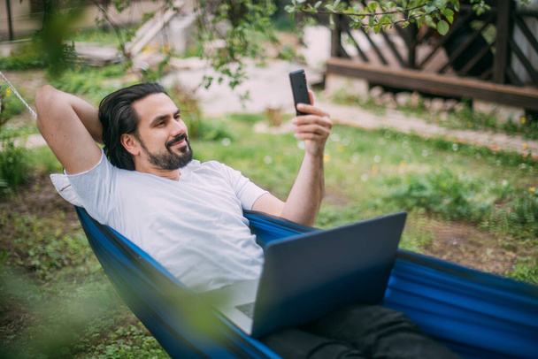 Un hombre trabaja con un portátil y un teléfono en una hamaca en una casa de campo. Un joven se encuentra en una hamaca con un ordenador, se comunica en línea, aprende de forma remota en la casa de campo en el verano - Foto, imagen