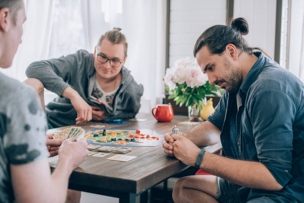 友人はリビングルームでボードゲームをプレイします。若い男の会社は、テーブルに座って、感情的かつ陽気に週末にカードゲームをプレイ - 写真・画像