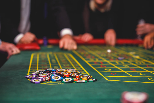 A close-up vibrant image of multicolored casino table with roulette in motion, with casino chips. the hand of croupier, mone and a group of gambling rich wealthy people in the backgroun - Zdjęcie, obraz