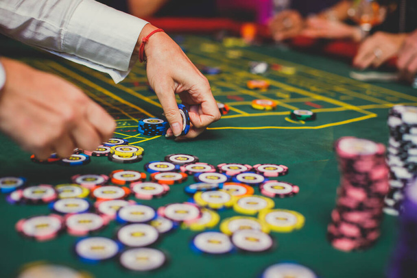 A close-up vibrant image of multicolored casino table with roulette in motion, with casino chips. the hand of croupier, mone and a group of gambling rich wealthy people in the backgroun - Фото, изображение