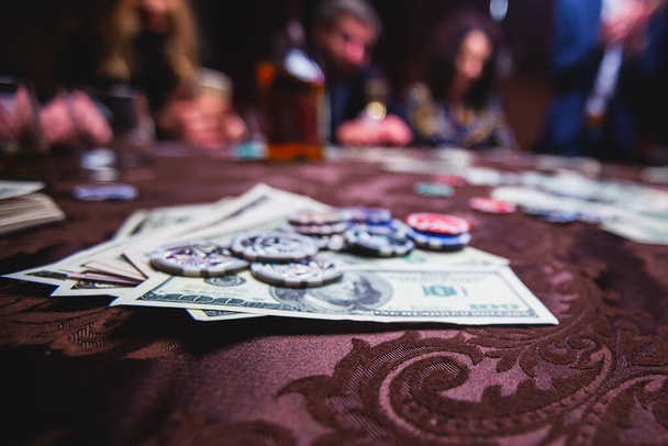 View of poker table with pack of cards, tokens, alcohol drinks, dollar money and group of gambling rich wealthy people playing poker - Foto, Bild