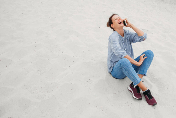 Jeune femme souriante parlant par téléphone sur une plage. Femme avec téléphone assis sur le sable blanc en arrière-plan. Belle fille a une conversation téléphonique joyeuse, rit et est heureux. - Photo, image