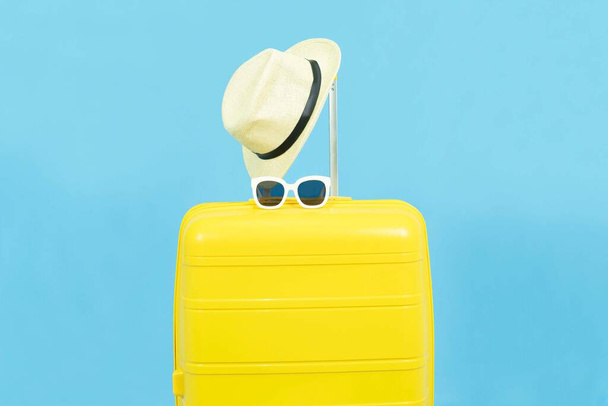 Summer holidays, vacation and travel concept.Yellow Suitcase or luggage bag and sunglasses on sun hat isolated on blue background - Фото, изображение