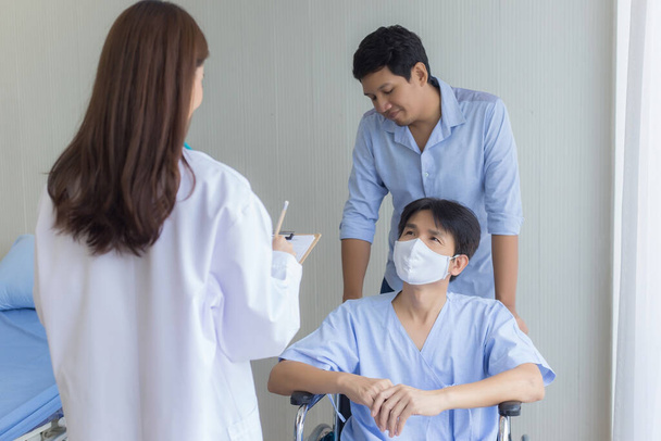 Aasialainen naislääkäri tarkistaa ja puhuu miespotilaan kanssa hänen oireestaan sairaalassa. Hän pukeutuu leikkausnaamariin ja istuu pyörätuolilla, kun toinen työntää sitä.. - Valokuva, kuva