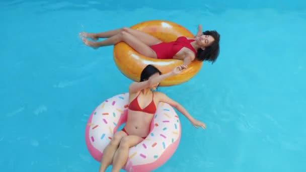 Two adult step sister swimming in family parents home pool holding hands and spinning. Couple mixed race woman in red swimsuits sitting in inflatable circle tube playing dancing tanning in blue water - Footage, Video