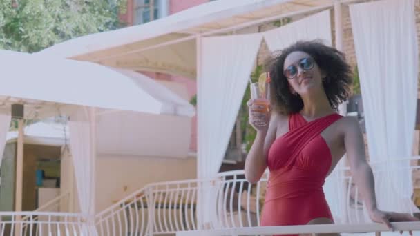 ヴィラスパホテルのプールサイドのテラスバルコニーにある流行の水着スタンドで誘惑的なモデルは、ノンアルコールカクテルを飲み、夏休みを祝い、挨拶をし、ガラスを上げ、カメラで浮気笑顔 - 映像、動画