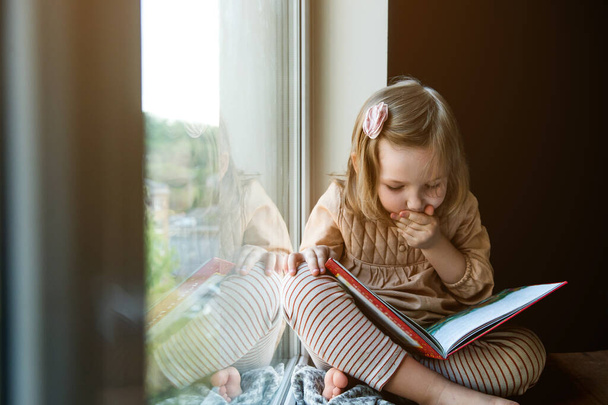 Pikkutyttö luki kirjan ikkunan lähellä. Koulutuskonsepti. Poika, jolla on kirja käsissään. Nopea lukea - Valokuva, kuva