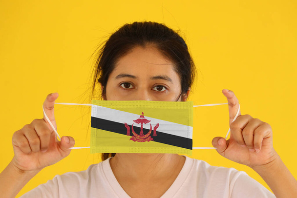 Eine Frau in weißem Hemd mit Brunei-Flagge auf Hygienemaske in der Hand und hochgezogenem Gesicht auf gelbem Hintergrund. Winzige Partikel oder Viren Corona oder Covid 19 Schutz. Konzept zur Bekämpfung von Krankheiten. - Foto, Bild