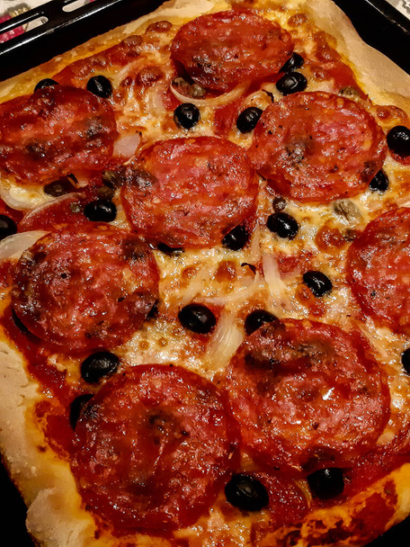 levensecht beeld van een zelfgemaakte pizza met pittige salami, zwarte olijven, tomaten en mozzarella - Foto, afbeelding