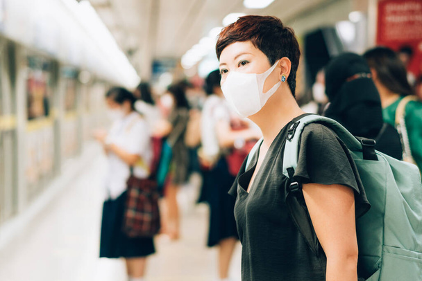 Piękna azjatycka podróżniczka w średnim wieku o zmartwionym wyglądzie, nosząca medyczną maskę na twarz w celu ochrony przed zakażeniem wirusami, pandemią, wybuchem i epidemią choroby w zatłoczonym podziemnym pociągu - Zdjęcie, obraz