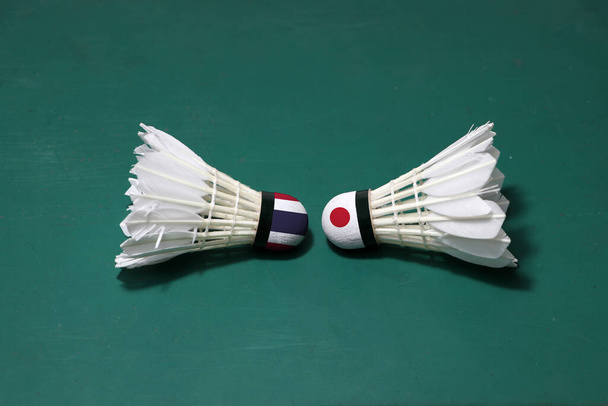 Twee gebruikte shuttlecocks op de groene vloer van Badminton Court met beide hoofden tegen elkaar. Een hoofd geschilderd met Thaise vlag en een hoofd geschilderd met de Japanse vlag, concept van badminton concurrentie tussen de twee landen. - Foto, afbeelding