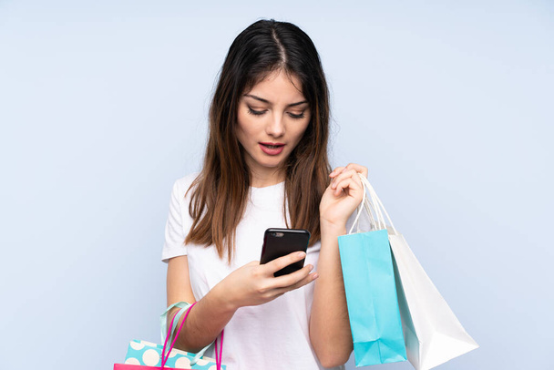 Młoda brunetka nad odizolowanym niebieskim tle trzymając torby na zakupy i pisząc wiadomość z telefonu komórkowego do znajomego - Zdjęcie, obraz