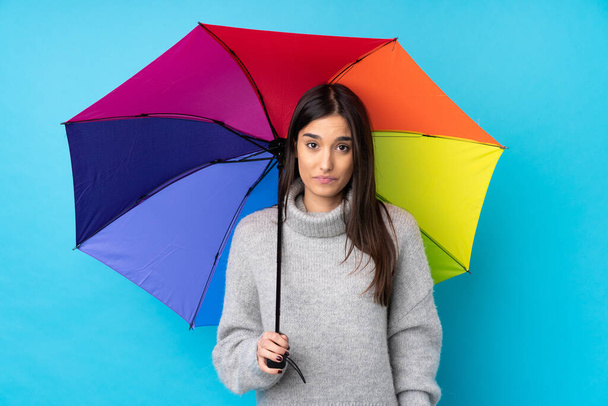 Young brunette woman holding an umbrella over isolated blue wall pleading - Foto, Bild